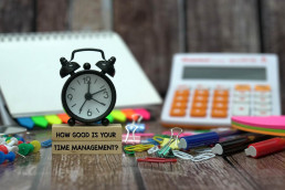 Small clock with 4 hands on top wooden blocks that spell out 'How good is your time management?', calculator and highlighter pens in the background